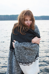 Young redhead woman wrapped in plaid blanket smiling, standing near lake. Autumn cold windy cloudy...