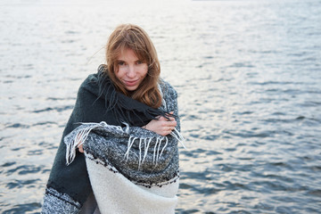 Young redhead woman wrapped in plaid blanket smiling, standing near lake. Autumn cold windy cloudy...