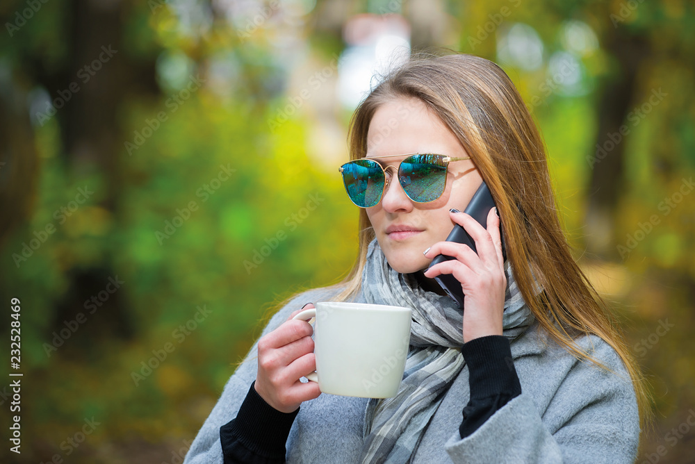 Wall mural beautiful young woman talking smartphone walking colorful autumn street. girl in a gray coat and sun