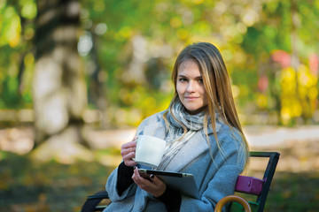 Young blonde girl with umbrella sitting on a park bench in the autumn season. girl in a gray coat drinks coffee, looks into a tablet, talks on the phone, reads a book, writes in a notebook