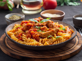 fusilli pasta with tomato sauce, chicken fillet with basil leaves on dark brown background, side...