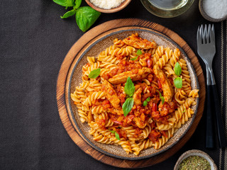 fusilli pasta with tomato sauce, chicken fillet with basil leaves on dark brown background, top view, copy space