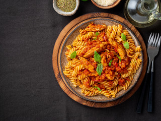 fusilli pasta with tomato sauce, chicken fillet with basil leaves on dark brown background, top view, copy space