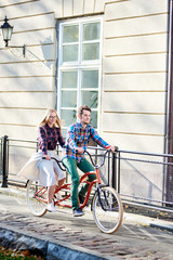 Young active smiling traveler couple, handsome bearded man and pretty blond woman in glasses riding together tandem bicycle along paved sidewalk on bright sunny autumn day by ancient buildings.