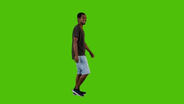 Casually dressed young man walking, waving hello and looking around happily in a full side shot over a green background.