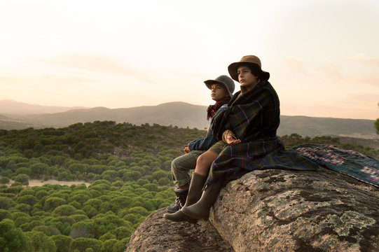 Two boys sitting on a stone covered with a blanket watching the sunset