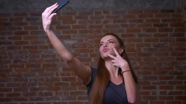 Amazing caucasian ginger female is showing her happiness while taking selfie pictures on web camera of her smartphone while standing, being relaxed, chilling in brick studio