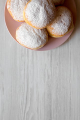 Homemade sweet donuts with powdered sugar on pink plate on white wooden background, overhead view. Copy space.