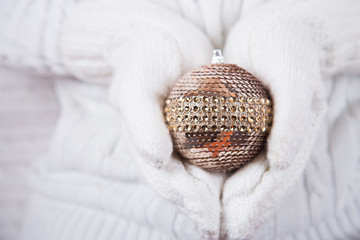 Hands in white knitted mittens holding glowing metallic Christmas ball.