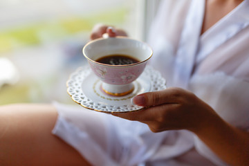 Girl in a Bathrobe sits on the windowsill next to the window and drinks coffee from a mug with a saucer. Young woman in a bathrobe drinking hot espresso or cappuccino in the morning on the windowsill.