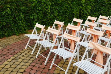White wooden chairs decorated with orange bows and roses for holiday or anniversary ceremony in row outdoors on green grass background. Wedding and celebration