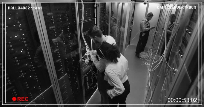 Wide monochrome shot from surveillance camera of people working in a data center server room with rows of server racks checking the equipment and discussing their work.