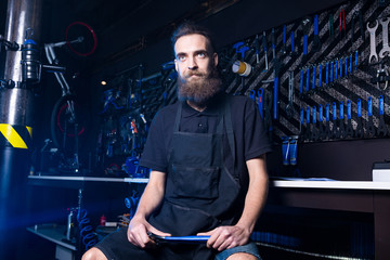 Obraz na płótnie Canvas Portrait of small business owner of young man with beard. Guy bicycle mechanic workshop worker sitting with tool in his hand in a working black clothes in an apron in the background of a bicycle shop