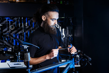 Theme sale and repair of bicycles. Young and stylish with a beard and long hair, a Caucasian man uses a tool to set up and repair a bike in a store. Business owner at work
