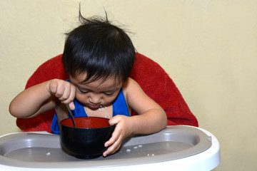 One 1 year old Asian baby boy learning to eat by himself, messy on baby dining chair at home