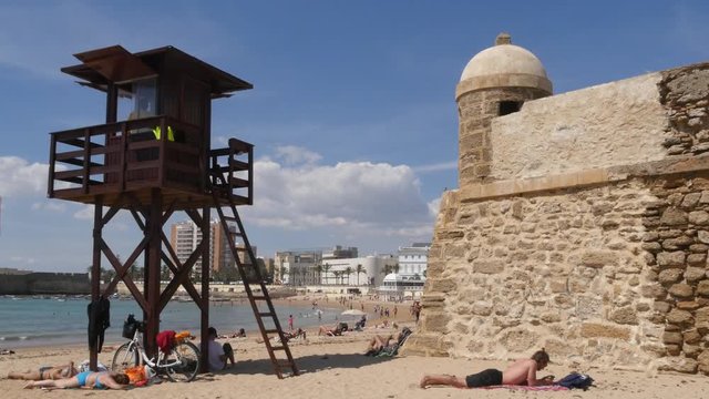Spanish City of Cadiz waterfront.
Camera Shot - Static shot of people sun bathing on La Caleta beach with Santa Catalina in the background.