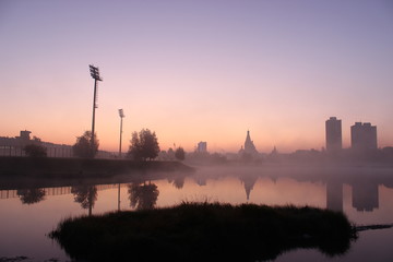 Fototapeta premium pink dawn at the misty lake. colored sky around. City background. skyscrapers around the old building