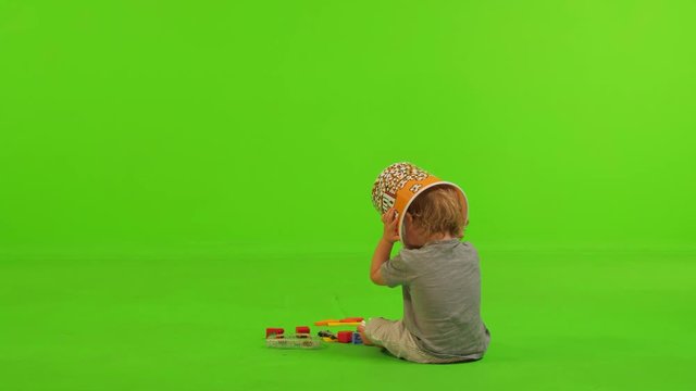 A toddler sits and plays peekaboo with himself with a popcorn bawl and laughs, over a green screen.