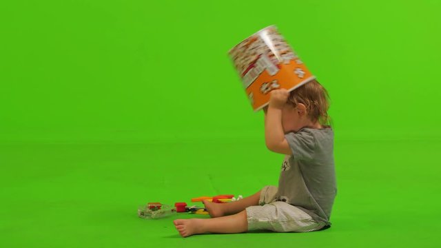 A toddler sits, plays and walk with a popcorn bawl  and some toys over a green screen.