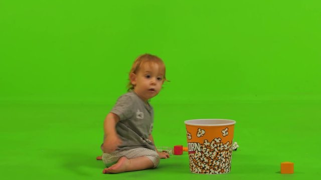 A toddler sits and happily put some toys inside a popcorn bawl over a green screen.