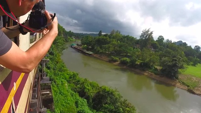 Tourist take pictures from old train windows. Train runs over the stip hanging old wooden bridge on Kwai River in Thailand