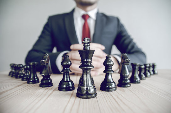 Retro Style Image Of A Businessman With Clasped Hands Planning Strategy With Chess Figures On An Old Wooden Table.