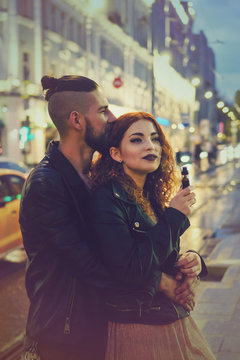 Couple in love on date. Boyfriend hugs and kisses girl from behind. She smokes an electronic cigarette. They walk streets of night city. Wet asphalt after rain.