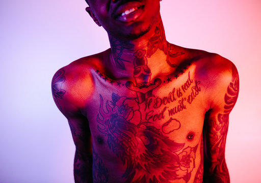 Bare chested heavily tattooed mans torso under red, pink and blue lighting