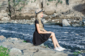 Fototapeta na wymiar Beautiful young woman blonde with long hair in a hat sitting on a rocky shore by the river and relaxing. Around the mountains and rocky landscape.