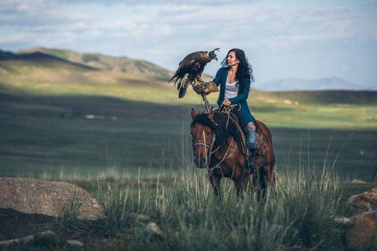 black haired woman riding through kyrgyz steppe with golden eagle on her arm