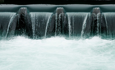close up of a manmade concrete weir in a city with a blue river cascading over it