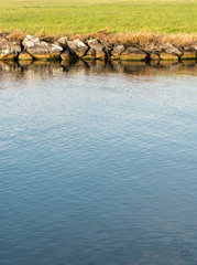 vertical view of a river canal with deep blue water and a rocky edge and green grass behind abstract view