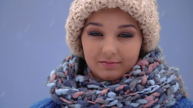 Happy calm women facing camera during winter holidays outside. Snow falling in slow motion 4K. Young gentle female enjoying winter day outdoors. Woman in cap, coat, scarf standing against blue copy