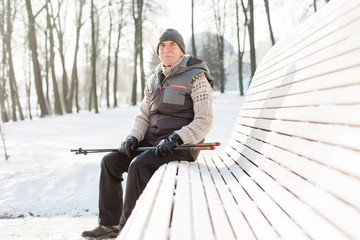 Senior man sitting with nordic walking poles in winter  park. Healthy lifestyle concept. Mature man resting after exercise outdoors.