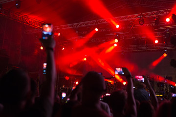a crowd of people lit by floodlights with red light, stand and shoot a music concert on the phone, blurred background