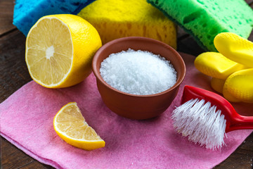 Fototapeta na wymiar Lemon acid in a small plate, slice of lemon, a juicy lemon, sponge for washing dishes, brushes and yellow rubber gloves for cleaning the house on wooden background. Natural cleaning products