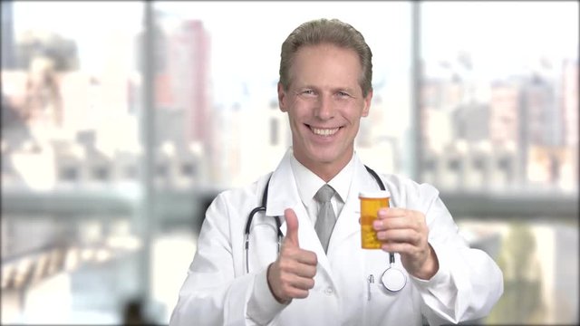 Positive male doctor, blurred background. Cheerful middle aged doctor with pill bottle on blurred background. Mature doctor with tablets gesturing thumb up. Ways of treatment.