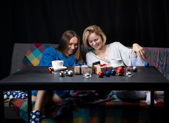 Women sit on the sofa in home clothes. On the table are cups, candles, New Year's toys and gifts. Black background.