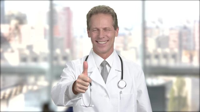 Handsome doctor gesturing thumb up. Cheerful middle-aged doctor showing thumb up gesture on window city background.