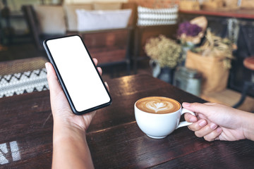 Mockup image of hands holding black mobile phone with blank screen while drinking coffee in vintage cafe