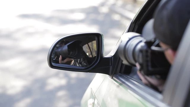 Reflection in side mirror of young private detective man sitting inside car and photographing with dslr camera