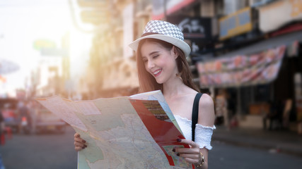 Cheerful woman wanderer with trendy look searching direction on location map in city centre. Happy female traveler and tourist concept,  Khao San Road walking street at Bangkok, Thailand