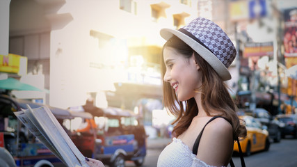 Cheerful woman wanderer with trendy look searching direction on location map in city centre. Happy female traveler and tourist concept,  Khao San Road walking street at Bangkok, Thailand