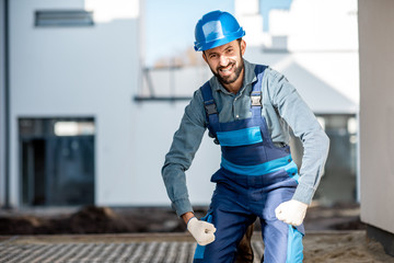 Funny portrait of a strong builder in uniform on the construction site with white houses on the background