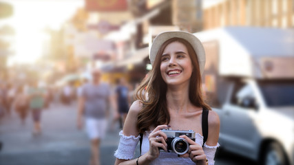 Beautiful young caucasian taking photos in city centre are walking Khaosan Road walking street in evening at Bangkok, Thailand. Happy female traveler and tourist concept.