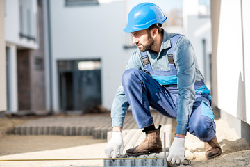 Builder in uniform mounting paving tiles on the construction site with white houses on the...