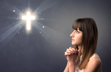 Young woman praying on a grey background with a shiny cross above her
