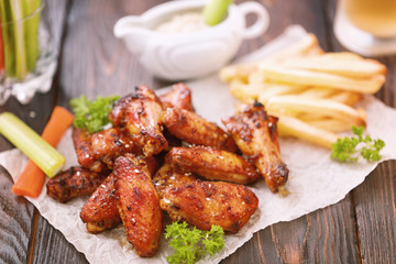 Roasted chicken wings served on a wooden table with glass of beer, french fries, carrot, celery and sause