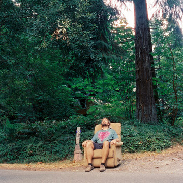 Bearded man in shorts and tie-dyed t-shirt relaxes in an easy chair on the side of a road.