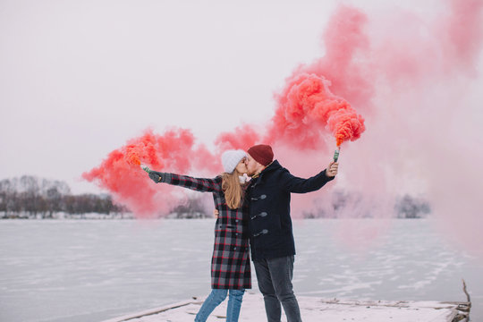 Young lovely couple having fun with red smoke grenades on the pier by frozen river